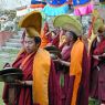 A procession of monks wearing yellow and red hats and carrying cymbals, religious texts, and parasols.